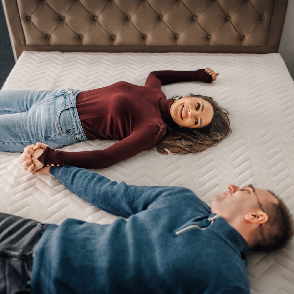Happy couple relaxing on a new bed, trying out its comfort and quality while shopping for furniture in a store